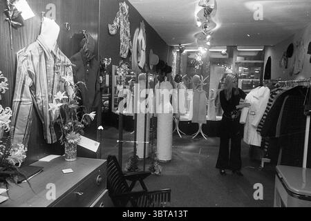 Magasin de mode intérieur pour femmes, Whizgle News, Dutch Desk, pays-Bas, 1950 - 2000 sur 12-1968. Ces rubriques sont illustrées dans l'image. La scène se déroule dans une boutique, présentant une variété d'articles de mode dans un espace confortable et bien organisé. D'un côté, les mannequins présentent des tenues élégantes, dont une robe glamour qui scintille sous le doux éclairage par le plafond. La palette de couleurs comprend un mélange de tons sourds et de teintes plus vibrantes, créant une atmosphère accueillante. Une femme, vêtue d'une élégante tenue noire avec un pantalon évasé et un haut ajusté, se tient près du centre de la pièce, examinez soigneusement Banque D'Images