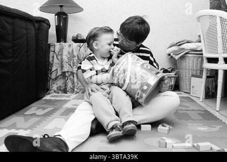 Soins à domicile chez Matz Gerritsen, Whizgle News, Dutch Desk, pays-Bas, 1950 - 2000 le 23-04-1995. L'image inclut ces rubriques. Dans un cadre intérieur cosy, une scène joyeuse se déroule avec un enfant et un adulte. La pièce est doucement éclairée, probablement par une lampe à proximité, qui projette une lueur chaude. L'atmosphère est remplie d'un sentiment de chaleur et d'affection, souligné par l'interaction étroite entre les deux figures. L'enfant, assis sur le sol, a les cheveux de couleur claire et est habillé d'une chemise rayée qui complète la nature ludique de l'environnement. Il est intentionnellement engagé avec une couleur Banque D'Images