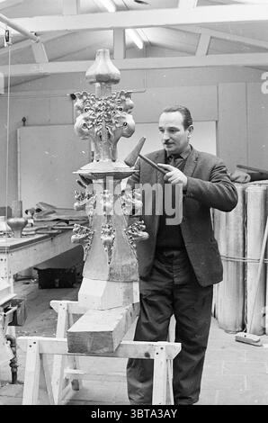 Travail sur l'oignon de la Grote ou Sint-Bavokerk tapant les spinnacles avec le plomb Grote Kerk Haarlem églises produit Bavokerk travail Haarlem Grote Markt pays-Bas, Whizgle News, Dutch Desk, pays-Bas, 1950 - 2000 on 02-02-1965. L'image inclut ces rubriques. Dans un cadre d’atelier rempli de lumière naturelle, un homme se tient concentré sur une structure décorative en bois qu’il fabrique. Il est habillé d'un costume sombre avec une veste ajustée et un pull en dessous, dégageant un sens du professionnalisme. Son expression est une expression de concentration car il tient un outil de sculpture dans une main, prêt à faire un Precis Banque D'Images