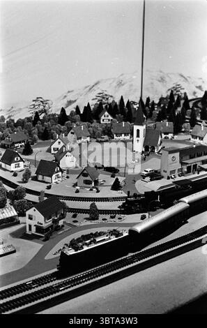 Train électrique à Joh. Des Grecs Birdsong Jeux et jouets, Whizgle News, Dutch Desk, pays-Bas, 1950 - 2000 le 06-07-1962. L'image inclut ces rubriques. La scène capture un paysage modèle miniature, riche en détails et caractérisé par un sentiment de nostalgie. À l'avant-garde, un train modèle épuré serpente le long d'une piste circulaire, scintillant légèrement sous les lumières environnantes. L’extérieur brillant du train contraste avec les tons discrets du paysage environnant. Sur fond de collines douces et vallonnées et de montagnes imminentes, le ciel est d'une ombre pâle, presque délavé à l'Enhanc Banque D'Images