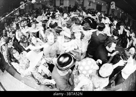 Carnival in Noordwijkerhout Carnival, Whizgle News, Dutch Desk, pays-Bas, 1950 - 2000 le 22-02-1971. Ces rubriques sont illustrées dans l'image. La scène capture un rassemblement intérieur animé grouillant d'énergie et de mouvement. Un groupe diversifié de personnes remplit l'espace, rayonnant une atmosphère de célébration et de camaraderie. Les individus sont habillés dans des tenues éclectiques vibrantes, caractérisées par des motifs audacieux et des couleurs vives. Certains portent des chapeaux et des accessoires amusants et surdimensionnés, rehaussant l'ambiance festive. L'éclairage est adouci et légèrement atténué, avec de petites ampoules enfilées à travers le plafond, créant Banque D'Images