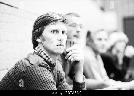 Portrait entraîneur de football en salle partenaire du maillot féminin : TEN Cate BV Agence de distribution Alkmaar, Whizgle News, Dutch Desk, pays-Bas, 1950 - 2000 sur 10-1980. Ces rubriques sont illustrées dans l'image. Dans un cadre monochrome, un groupe d'hommes est engagé dans la conversation. L'accent est mis sur un individu avec des caractéristiques distinctes, y compris des cheveux de longueur moyenne, légèrement touffus et une expression réfléchie. Il arbore un pull boutonné superposé sur une chemise à col, transmettant un sentiment de style décontracté mais délibéré. Sa main est levée à son menton, suggérant la contemplation ou la curiosité. Derrière lui, deux OTH Banque D'Images