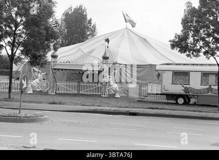 Circus Bongo Schalkwijk Haarlem pays-Bas, Whizgle News, Dutch Desk, pays-Bas, 1950 - 2000 le 10-06-1991. Voici les rubriques de l'image. La scène présente une grande tente de cirque, caractérisée par son auvent rayé audacieux dans des couleurs alternées de rouge et de blanc. La tente est bien en vue sur une tache ouverte d'herbe, entourée d'une clôture de piquet blanche basse qui ajoute une touche pittoresque au cadre. Flanquant la tente sont deux figures de clown fantaisistes, peintes de couleurs vives, prêtes dans des poses ludiques. Ils servent de décorations accrocheuses, rehaussant l'atmosphère festive. À l'avant Banque D'Images