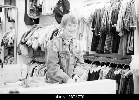 À la mode pour enfants Haarlem pays-Bas, Whizgle News, Dutch Desk, pays-Bas, 1950 - 2000 le 09-02-1993. L'image montre ces rubriques. Dans un magasin de vêtements animé, la scène met en valeur une atmosphère chaleureuse et accueillante. Une personne se tient derrière un comptoir blanc, souriant joyeusement, créant une ambiance accessible. L'individu a les cheveux courts, de couleur claire et est habillé d'une veste en denim classique, qui ajoute une touche décontractée au cadre. Autour de la figure est un affichage vibrant de porte-vêtements débordant de divers vêtements. Les tissus sont variés, avec des textures et Banque D'Images