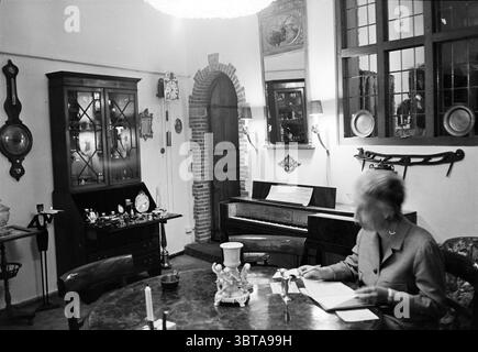 Antiquaire Jacobijnestraat Interior Haarlem Jacobijnestraat pays-Bas, Whizgle News, Dutch Desk, pays-Bas, 1950 - 2000 le 19-08-1964. Voici les rubriques de l'image. La scène dispose d'une salle confortable et accueillante remplie d'un éventail de meubles et de décoration soigneusement agencés. Au centre, une table ronde à la surface sombre est entourée de quelques chaises en bois. Une chaise est notablement occupée par une femme âgée, qui est engagée dans l'écriture ou la lecture de quelque chose avec une expression focalisée, bien que son visage soit flou. À gauche, il y a un grand meuble orné en bois sombre wi Banque D'Images