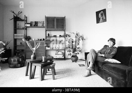 Intérieur de la maison de MRS Barendregt à Brinkmann Complex Brinkman passage personnes Haarlem Grote Markt pays-Bas, Whizgle News, Dutch Desk, pays-Bas, 1950 - 2000 on 03-03-1982. L'image contient ces rubriques. La scène représente un salon confortable caractérisé par une atmosphère chaleureuse et accueillante. Au centre de l'espace se trouve un canapé profond de couleur sombre, orné de coussins détendus. Une personne est assise confortablement sur le canapé, portant une tenue aux tons clairs et affichant un sourire paisible, suggérant une humeur de contenu. A gauche, une petite table en bois sombre tient un modeste arrangeme Banque D'Images