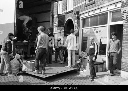 Camion de chargement pour Poland transport, Whizgle News, Dutch Desk, pays-Bas, 1950 - 2000 sur 18-04-1985. Ces rubriques apparaissent dans l'image. Dans un cadre urbain animé, un groupe d'individus est engagé dans une scène active à l'extérieur d'un magasin caractérisé par de grandes fenêtres ornées d'un décor simple. L'atmosphère transmet un sentiment de transition ou de mouvement, impliquant probablement une livraison ou une opération de déchargement. La scène montre un camion ouvert à l'arrière, révélant des gens à l'intérieur qui sont occupés à trier ou distribuer des articles. Ils portent des vêtements décontractés, y compris des chandails et des jeans, ce qui indique un relax Banque D'Images