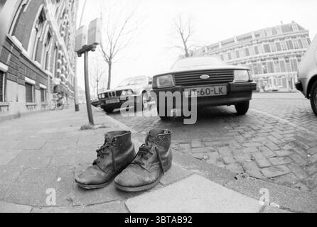 Paire de chaussures à côté du parcmètre Nassauplein Chaussures Haarlem Nassauplein pays-Bas, Whizgle News, Dutch Desk, pays-Bas, 1950 - 2000 le 09-01-1986. L'image contient ces rubriques. Dans un environnement urbain animé, la scène se déroule sur une rue pavée, où la texture du trottoir ajoute un charme rustique. Deux voitures stationnées, l'une un modèle classique avec un design élégant et angulaire au premier plan, et l'autre une berline plus moderne, sont côte à côte, leurs couleurs atténuées par la palette monochrome. Les voitures sont juxtaposées contre les briques complexes des bâtiments voisins, caractérisés par Banque D'Images
