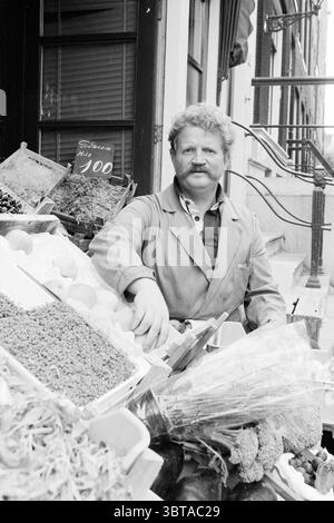 Toon Labes Vegetable Man A'dam (radio) Vegetables, Whizgle News, Dutch Desk, pays-Bas, 1950 - 2000 le 01-08-1978. Ce sont les éléments de l'image. La scène présente un cadre de marché extérieur animé, vibrant avec la présence de produits frais et divers articles à vendre. Au premier plan, un vendeur d'âge moyen avec une moustache distinctive et touffue se trouve confortablement à côté d'une gamme de fruits et légumes colorés. Il porte un tablier de couleur claire sur une chemise à manches longues, suggérant un comportement pratique mais accessible. Autour de lui sont soigneusement disposés des caisses et des bacs remplis d'un V. Banque D'Images