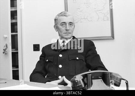 Mr. Brands chef de la police de la salle des personnes police, Whizgle News, Dutch Desk, pays-Bas, 1950 - 2000 on 20-02-1970. Ces rubriques sont illustrées dans l'image. La scène montre un homme assis à un bureau, vêtu d'un uniforme sombre de style militaire qui suggère un rôle autoritaire. Le tissu de l'uniforme est noir profond avec des boutons brillants, ajoutant une sensation de formalité. Ses cheveux sont soigneusement stylisés, mettant en valeur un mélange de nuances grises et foncées, mettant l'accent sur la maturité et l'expérience. Le cadre est un bureau intérieur, caractérisé par un agencement propre et épuré. Derrière lui, un mur est orné d'une carte Banque D'Images