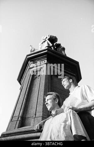 Deux joueurs de football jaune-blanc, Whizgle News, Dutch Desk, pays-Bas, 1950 - 2000 le 16-08-1993. Ce sont les éléments de l'image. La scène capture deux hommes debout devant un grand monument orné. Le monument, fabriqué en pierre sombre, présente des sculptures complexes et une statue à son sommet. La statue semble représenter une figure historique, avec une pose imposante et des caractéristiques détaillées, faisant allusion à la grandeur et à l'importance. Les hommes sont bien placés au premier plan, appuyés contre le monument. Un homme a une expression détendue, avec un léger sourire, vêtu d'un lig Banque D'Images