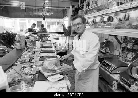 International Boucherie Broekhof Haarlem Kruisstraat pays-Bas, Whizgle News, Dutch Desk, pays-Bas, 1950 - 2000 on 06-1988. L'image inclut ces rubriques. La scène représente une charcuterie ou une boucherie animée, remplie d'un éventail de délices et d'une atmosphère animée. Au premier plan, un homme d'âge moyen portant un manteau blanc de chef et des verres coupe habilement un gros morceau de viande. Son expression focalisée suggère une expertise et une fierté dans son métier. Derrière lui, le comptoir est orné de diverses viandes et fromages, soigneusement disposés et exposés. La surface en marbre ajoute une touche d'élégance Banque D'Images
