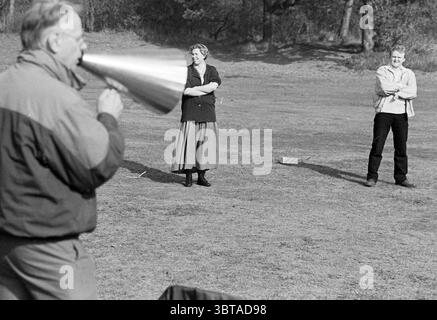 Cours de gestion sur un terrain avec corne, Whizgle News, Dutch Desk, pays-Bas, 1950 - 2000 le 04-1995. Ces rubriques apparaissent dans l'image. Dans un espace ouvert herbeux, trois personnes participent à une scène imprégnée d'un sentiment de communauté et d'engagement. Au premier plan, un homme aux cheveux gris utilise un grand mégaphone métallique pour projeter sa voix. Il porte une veste décontractée et apparaît animé, projetant l'excitation ou l'urgence. À quelques pas se trouvent deux personnes qui semblent écouter attentivement. Une femme, avec une coiffure légère et une blouse à motifs, a les bras croisés, Banque D'Images