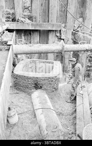 Ouvrages d'assainissement Parklaan Works Haarlem Parklaan pays-Bas, Whizgle News, Dutch Desk, pays-Bas, 1950 - 2000 on 15-12-1965. L'image inclut ces rubriques. La scène capture un chantier de construction caractérisé par une esthétique industrielle terreuse. Au centre de la composition se trouve une structure circulaire en briques qui semble être un puits ou une fondation, élaborée avec des briques rouges et brunes. Autour de la structure se trouvent des tas de sable et de saleté, suggérant une excavation en cours. A gauche, un tuyau industriel s'étend horizontalement à travers la scène, sa surface métallique altérée contraste Banque D'Images