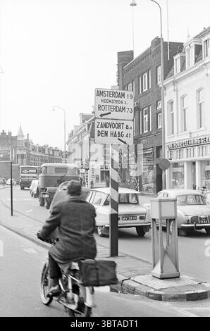 Panneau avec flèches Houtplein Traffic Safe Traffic Haarlem Houtplein pays-Bas, Whizgle News, Dutch Desk, pays-Bas, 1950 - 2000 le 17-03-1965. Ce sont les éléments de l'image. La scène représente une rue urbaine animée, caractérisée par un mélange d'architecture historique et du milieu du XXe siècle. Les bâtiments bordant la rue vont de façades en briques à des designs plus contemporains, avec un mélange de devantures de magasins et d'espaces résidentiels. Au premier plan, un cycliste vêtu d'un long manteau sombre navigue devant une rangée de voitures anciennes garées, leurs formes douces et arrondies indicatives de l'époque. Le bicyc Banque D'Images