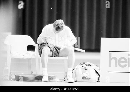 Match de tennis masculin, Whizgle News, Dutch Desk, pays-Bas, 1950 - 2000 le 09-04-1989. L'image contient ces rubriques. Dans la scène, un athlète est assis sur une simple chaise en plastique blanc, paraissant concentré et quelque peu contemplatif. Vêtu d'un maillot de sport de couleur claire et d'un short, l'individu se penche légèrement en avant, examinant méticuleusement une raquette de tennis dans ses mains. Le cadre suggère une installation sportive intérieure, caractérisée par l'éclairage tamisé au-dessus qui crée un contraste avec les zones plus lumineuses du court. L'arrière-plan présente un rideau sombre et drapé, améliorant l'inti Banque D'Images