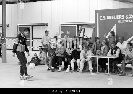 Gert Jan Oudshoorn maintenant le record du monde Hillegom tentatives et améliorations de records Hillegom, Whizgle News, Dutch Desk, pays-Bas, 1950 - 2000 le 25-07-1987. Voici les rubriques de l'image. Dans un vaste cadre industriel, un groupe de spectateurs est assis le long d'un côté, regardant attentivement un jeune démontrant des compétences avec un ballon de football. Le fond présente un mur partiellement peint et une rangée de camions garés à proximité, ajoutant un élément de profondeur à la scène. Les spectateurs, un mélange diversifié d'âges, présentent différentes expressions allant de la concentration à l'amusement. Banque D'Images