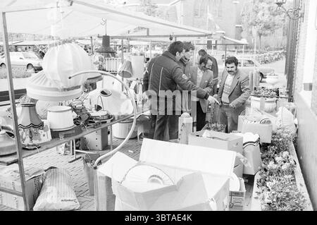 Marché aux puces Hillegom marché Hillegom, Whizgle News, Dutch Desk, pays-Bas, 1950 - 2000 le 12-05-1984. Ces rubriques apparaissent dans l'image. La scène capture un marché en plein air animé rempli de divers vendeurs et acheteurs. Au premier plan, plusieurs caisses et boîtes sont empilées de manière désordonnée, contenant un éventail d'objets ménagers, principalement des abat-jours et des objets décoratifs. Les couleurs des produits vont des tons neutres aux teintes vives et accrocheuses, ajoutant de l'énergie à l'environnement. Quelques individus sont rassemblés, engagés dans la conversation, leurs postures suggérant un Li Banque D'Images
