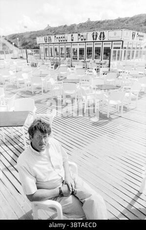 Propriétaire Freddy et Paul Meerendonk Cafe Beach and Beaches, Whizgle News, Dutch Desk, pays-Bas, 1950 - 2000 on 31-05-1989. L'image inclut ces rubriques. La scène capture un cadre extérieur tranquille, avec une vaste terrasse en bois qui s'étend vers une série de chaises blanches et de tables arrangées soigneusement. L'atmosphère suggère un endroit côtier, probablement près d'une plage, comme indiqué par la configuration décontractée et détendue. Au premier plan, un homme d'âge moyen s'assoit confortablement sur une chaise en plastique blanc, portant une chemise à manches courtes de couleur claire. Son expression est contemplative, peut-être réfléchie, Banque D'Images