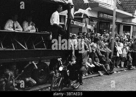 Anneau couture sur un cyclomoteur jeux de vacances jours de vacances camp de vacances, Whizgle News, Dutch Desk, pays-Bas, 1950 - 2000 le 03-08-1971. L'image inclut ces rubriques. Dans une scène urbaine animée, une foule animée se rassemble le long d'une rue étroite, regardant avec impatience un événement se dérouler. L'atmosphère est vibrante et remplie d'anticipation. Au premier plan, un jeune homme fait du vélo, croisant en toute confiance devant les spectateurs. Il porte un costume, ce qui contraste avec la tenue décontractée de ceux qui l'entourent, faisant allusion à un charme ludique. Son expression est concentrée, peut-être fière de sa présence au milieu Banque D'Images