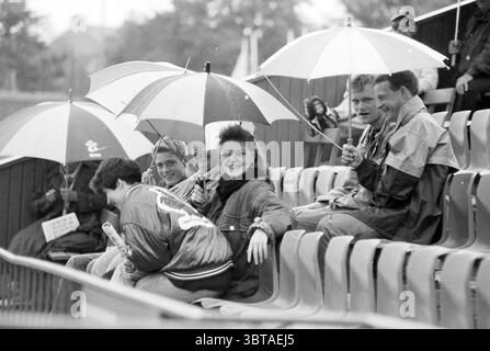 Baseball Nicols - Spartan, Whizgle News, Dutch Desk, pays-Bas, 1950 - 2000 on 29-06-1991. Ce sont les éléments de l'image. Dans un cadre extérieur animé, plusieurs personnes sont rassemblées dans une série de sièges de style gradin, protégés de la bruine par des parapluies colorés. L’ambiance est animée malgré la pluie, avec un sentiment de camaraderie au sein du groupe. Les gens montrent une gamme d'expressions, des sourires joyeux à la conversation informelle. Parmi eux, un individu se distingue par une coiffure distinctive et une posture détendue, indiquant le confort dans leur environnement. L'autre Banque D'Images