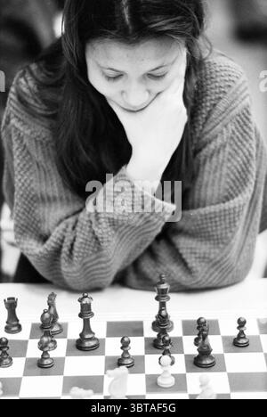 Tournoi d'échecs de Hoogovens : four-Way matches, Whizgle News, Dutch Desk, pays-Bas, 1950 - 2000 sur 20-01-1996. Ces rubriques sont illustrées dans l'image. Dans cette scène, une jeune femme est profondément concentrée sur un jeu d'échecs, son menton reposant soigneusement sur ses mains. L'expression sur son visage reflète la concentration et la détermination, incarnant l'état d'esprit d'un stratège. L'échiquier est l'élément central, avec un mélange de pièces de bois sombres et clairs, certaines élégamment sculptées, positionnées dans une disposition tendue en milieu de jeu qui laisse entrevoir une bataille complexe de l'esprit. Les couleurs contrastées des pièces d'échecs Banque D'Images