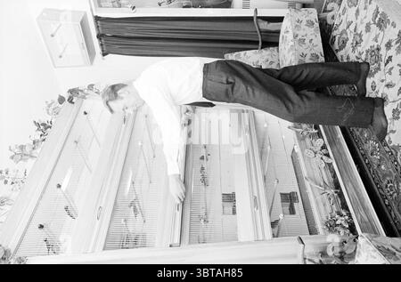 Pile de cages à oiseaux dans le salon volière appartement., Whizgle News, Dutch Desk, pays-Bas, 1950 - 2000. L'image inclut ces rubriques. Dans un cadre intérieur lumineux et confortable, un homme est capturé dans un moment de concentration alors qu'il interagit avec une grande armoire en bois qui dispose de portes vitrées. L'armoire présente une variété d'articles décoratifs, y compris éventuellement des plantes, des livres ou des objets de collection, qui sont partiellement masqués par les portes. L'homme, vêtu d'une chemise blanche et nette et d'un pantalon sombre, semble fiancé alors qu'il ouvre l'une des portes de l'armoire. Sa posture penche légèrement en avant, moi Banque D'Images