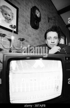 Jeroen Westerhuis roue de la Fortune, Whizgle News, Dutch Desk, pays-Bas, 1950 - 2000 le 02-04-1991. Ces rubriques sont illustrées dans l'image. La scène capture un intérieur confortable mais légèrement nostalgique. Un homme est assis sur un canapé sombre, penché en avant, les bras croisés, regardant soigneusement le spectateur. Il a les cheveux courts et foncés et un léger sillon dans son front, suggérant un moment de réflexion ou de contemplation. L'éclairage est doux, créant une atmosphère chaleureuse qui contraste avec l'esthétique noir et blanc. Derrière lui, le mur est une brique texturée, qui ajoute de la profondeur et un charme rustique Banque D'Images