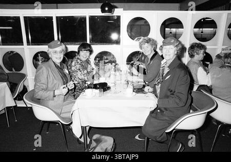 Réunion de 150 agents de bord KLM Reunion, Whizgle News, Dutch Desk, pays-Bas, 1950 - 2000 le 21-12-1985. Ces rubriques sont illustrées dans l'image. Dans une salle à manger moderne et bien éclairée, quatre femmes s'assoient autour d'une table drapée d'un tissu blanc éclatant. Le décor présente une toile de fond de grandes fenêtres circulaires, permettant une interaction dynamique de la lumière et de l'ombre. Chaque femme a un look unique : un mélange de vêtements élégants et d'accessoires élégants, des foulards aux chapeaux, créant un sentiment d'individualité tout en mettant en valeur des couleurs coordonnées, principalement dans des tons pastels doux et discrets. Les femmes sont fiancées moi Banque D'Images
