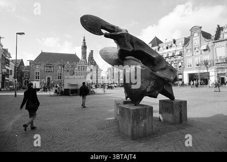 Sunfighter Gr. Marché Haarlem Grote Markt pays-Bas, Whizgle News, Dutch Desk, pays-Bas, 1950 - 2000 le 21-03-1991. Ce sont les éléments de l'image. Dans une place urbaine animée, une grande sculpture abstraite capte l'attention, ses formes organiques et ses surfaces texturées créant un point focal saisissant. La sculpture, composée de matériaux sombres et lourds, semble se tordre et s'étendre vers l'extérieur, comme si elle était en mouvement, contrastant fortement avec l'environnement environnant. A gauche, on voit un personnage marchant, leur silhouette définie contre les pavés texturés. Vêtu d'un manteau foncé, t Banque D'Images
