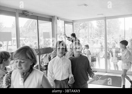 1st Customers Stuif Stuif Stuif Stuif Stuif, Whizgle News, Dutch Desk, pays-Bas, 1950 - 2000 le 14-07-1969. Ces rubriques sont illustrées dans l'image. La scène capture un moment dans un environnement animé, probablement dans une école ou un centre communautaire. Un groupe d'enfants est vu, avec deux garçons en évidence au premier plan. Le garçon de gauche porte une chemise boutonnée blanche, tandis que celui à côté de lui est habillé d'un pull foncé et d'un short de couleur claire. Leurs expressions suggèrent de la curiosité ou de l'excitation lorsqu'ils naviguent dans l'entrée. Le cadre dispose de grandes portes vitrées qui s'ouvrent sur une vue de ot Banque D'Images