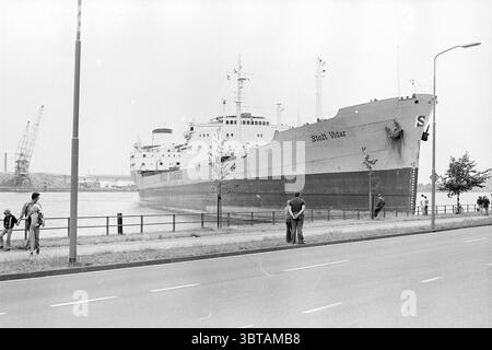 Pétrolier Noordersluis IJmuiden Ship collision, Whizgle News, Dutch Desk, pays-Bas, 1950 - 2000 le 13-07-1976. Ce sont les éléments de l'image. La scène montre un grand cargo amarré près d'un front de mer, dominant la composition avec sa structure imposante. La coque du navire, peinte dans des tons de gris, contraste avec le milieu environnant. Son nom, bien que non visible, fait allusion à son but et à ses liens avec l'industrie maritime. Au premier plan, deux individus se tiennent ensemble, engagés dans la conversation, tandis que quelques autres peuvent être vus plus loin sur le trottoir, soit marchant ou stoppi Banque D'Images