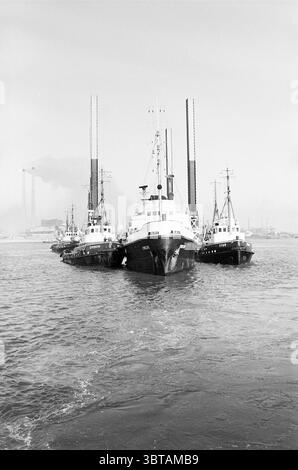Pièces de plate-forme de remorquage, Whizgle News, Dutch Desk, pays-Bas, 1950 - 2000 en 1967. Voici les rubriques de l'image. La scène présente un groupe de bateaux ancrés les uns aux autres sur l’eau, leurs coques peintes dans différentes nuances de noir et blanc, reflétant une esthétique maritime classique. Le navire central est plus grand, avec une structure solide et un gréement proéminent, tandis que deux bateaux plus petits le flanquant semblent plus profilés, ce qui suggère qu'ils sont conçus pour la maniabilité. Le ciel au-dessus de la tête est un gris sourd, créant une ambiance globalement sombre qui contraste avec la présence plus sombre de Banque D'Images