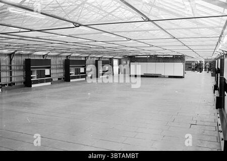 Mobilier de métro d'évacuation à Beverwijk évacuation, Whizgle News, Dutch Desk, pays-Bas, 1950 - 2000 le 03-04-1975. Voici les rubriques de l'image. La scène représente un intérieur spacieux, principalement monochrome, caractérisé par une disposition expansive et ouverte. Le sol est fait de grands carreaux de pierre rectangulaires, disposés en quadrillage, contribuant à un sentiment d'uniformité et de simplicité. Au-dessus, un toit transparent composé de structures en forme de grille permet à la lumière diffuse de filtrer à travers, projetant des ombres douces et créant une atmosphère lumineuse mais tamisée. Des deux côtés de l'espace, t Banque D'Images