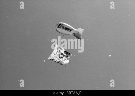 Montgolfière avec publicité pour l'entreprise de construction Vermeer, Whizgle News, Dutch Desk, pays-Bas, 1950 - 2000 en 1986. L'image contient ces rubriques. Dans la scène, un grand blimp plane gracieusement contre un ciel expansif, créant un point focal frappant au-dessus. Le blimp présente une forme profilée, avec une surface lisse qui reflète la lumière du soleil, soulignant sa flottabilité. Son extérieur présente un design audacieux et minimaliste caractérisé par le mot « VERMEER » sur le côté, faisant probablement référence à une marque ou à un événement. Sous le blimp, une bannière vibrante est suspendue, flutterin Banque D'Images