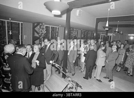 Jub. Réception Mr. de Graaf (Sports Affairs) Jubilee anniversaires, Whizgle News, Dutch Desk, pays-Bas, 1950 - 2000 le 01-10-1974. Ces rubriques apparaissent dans l'image. La scène capture un rassemblement dans un intérieur spacieux et moderne, caractérisé par des éléments de design du milieu du siècle. La chambre dispose d'un sol poli de couleur claire qui reflète l'éclairage artificiel doux fourni par les luminaires suspendus aux teintes arrondies. Les murs sont ornés de textures subtiles, complétées par des plantes stratégiquement placées qui ajoutent une touche de verdure à l'ambiance. Des groupes de personnes sont engagés dans conversati Banque D'Images