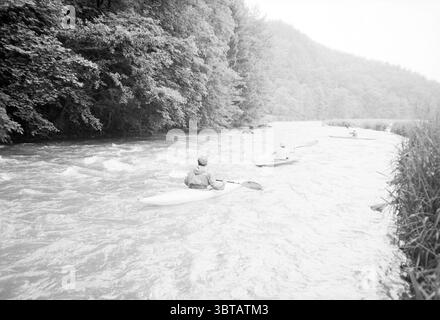 Survival tour belge. Ardennes Survival Tour, Whizgle News, Dutch Desk, pays-Bas, 1950 - 2000 le 30-06-1985. L'image contient ces rubriques. La scène se déroule le long d'une rivière qui coule doucement, entourée d'une végétation luxuriante. L'eau est un gris sourd, reflétant le ciel doux et nuageux au-dessus, créant une atmosphère sereine mais monochromatique. Un petit groupe d'individus pagaie dans des kayaks, leurs silhouettes contrastent avec la toile de fond plus claire de la rivière. Les kayaks sont de différentes nuances de blanc et de gris, se fondant parfaitement dans le cadre tranquille. La végétation flanquant la rivière se compose Banque D'Images