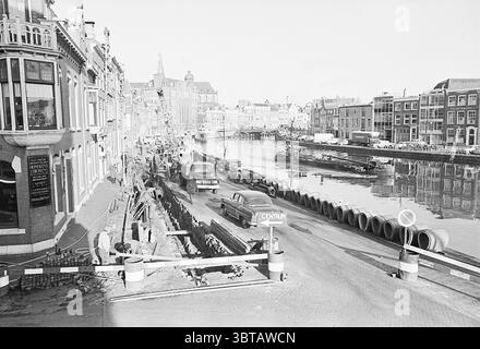Travaux sur les travaux de renouvellement des eaux usées de Spaarne, Whizgle News, Dutch Desk, pays-Bas, 1950 - 2000 le 29-01-1964. Ces rubriques apparaissent dans l'image. La scène représente une zone urbaine animée le long d'un canal serein, caractérisé par un mélange d'architecture historique et moderne. D'un côté, les bâtiments traditionnels aux façades ornées présentent des fenêtres décoratives et des toits à pignons, reflétant les styles architecturaux d'antan. Les murs sont ornés de teintes discrètes, principalement des nuances de beige et de brun clair, contribuant à une ambiance chaleureuse et nostalgique. Au premier plan, les travaux de construction sont en progres Banque D'Images
