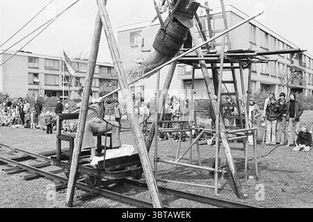 Ton.stenen Bloemendaal Queen's Day, Whizgle News, Dutch Desk, pays-Bas, 1950 - 2000 le 30-04-1977. L'image contient ces rubriques. La scène se déroule à l'extérieur, probablement dans une zone herbeuse entourée de bâtiments de faible hauteur, indiquant un environnement communautaire ou résidentiel. Une structure en bois, ressemblant à une grue de fortune ou à une catapulte, se tient bien en évidence au premier plan, construite à partir de poutres et de supports en bois brut. Une personne est activement engagée avec l'appareil, semblant tirer un levier ou manipuler un mécanisme qui est fixé à un grand seau. Ce seau est surélevé et semble l'être Banque D'Images