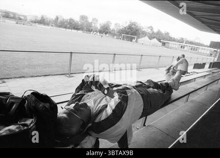 Entraîneur américain Atlanta, Whizgle News, Dutch Desk, pays-Bas, 1950 - 2000 sur 02-08-1992. Ces rubriques sont illustrées dans l'image. La scène capture un moment sur un terrain de sport, probablement un terrain de football, vu depuis un coin salon couvert. Au premier plan, deux individus occupent le banc. Une personne s'incline confortablement, vêtue d'une veste légère et d'un pantalon décontracté. Il a une posture détendue, peut-être se reposer après une longue journée. Sa tête est inclinée vers l'arrière, et des lunettes de soleil peuvent être vues perchées sur son visage, ajoutant une sensation de calme au cadre. A proximité, une autre figure est assise debout, engagée dans un plus actif Banque D'Images