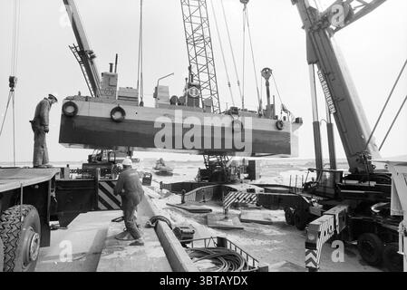 Dragueur de sable sur la jetée sud IJmuiden IJmuiden pays-Bas, Whizgle News, Dutch Desk, pays-Bas, 1950 - 2000 on 28-03-1993. Ce sont les éléments de l'image. La scène capture un environnement industriel occupé avec une palette monochrome, soulignant un sentiment de robustesse et de travail acharné. Au premier plan, deux ouvriers, vêtus d’un équipement de protection qui contraste fortement avec les tons sourds, sont engagés dans l’opération. Un ouvrier se tient à côté d'une énorme pièce de machinerie, son attention se concentrant sur la tâche à accomplir, tandis que l'autre semble en mouvement, coordonnant probablement le lifti Banque D'Images