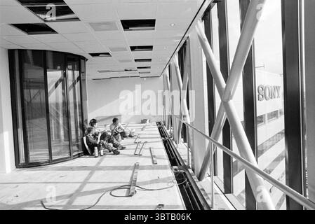 Intérieur Sony City in Badhoevedorp Badhoevedorp, Whizgle News, Dutch Desk, pays-Bas, 1950 - 2000 on 27-10-1992. Ce sont les éléments de l'image. La scène capture un intérieur spacieux en construction, caractérisé par une façade en verre expansive qui permet à la lumière naturelle d'inonder l'espace. Le revêtement de sol est inachevé, révélant des planches de bois brut, tandis que certaines zones semblent encombrées d'outils et d'équipements. Deux personnes, vêtues d'une tenue de travail, sont assises sur le sol, engagées dans une tâche qui implique l'assemblage ou la réparation de l'équipement. Ils sont concentrés, profondément engagés dans leur travail, s Banque D'Images