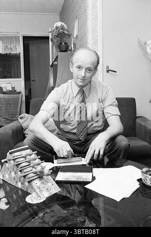 Mr de Groot avec toto Forms in Castricum Persons, Whizgle News, Dutch Desk, pays-Bas, 1950 - 2000 on 05-02-1974. L'image montre ces rubriques. Dans un cadre intérieur confortable, un homme est assis sur un canapé rembourré vert foncé, dégageant un comportement calme. Il a les cheveux de couleur claire, étroitement coupés, et porte une chemise claire soigneusement pressée avec une cravate à motifs qui présente des nuances de bleu, rouge et blanc. Sa posture est détendue mais attentive car il se penche légèrement vers l'avant, les mains posées juste au-dessus d'une petite table rectangulaire devant lui. Sur la table, une variété d'éléments créent une scène composée : une pile de Banque D'Images