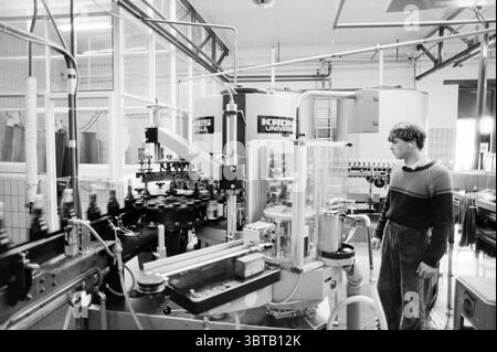 Nouveau fabricant de machines de remplissage de bouteilles Loots Drinks and Liquor Stores, Whizgle News, Dutch Desk, pays-Bas, 1950 - 2000 le 03-04-1985. Ces rubriques sont illustrées dans l'image. La scène représente un environnement industriel, caractérisé par un cadre d'usine qui met l'accent sur les machines et la production. Au centre de la composition se trouvent plusieurs grandes machines complexes, qui sont généralement métalliques avec une combinaison de finitions argentées et noires. Divers tubes et courroies transporteuses serpentent à travers les machines, faisant allusion à un système complexe de processus impliqués dans la fabrication. Au premier plan, un pe Banque D'Images