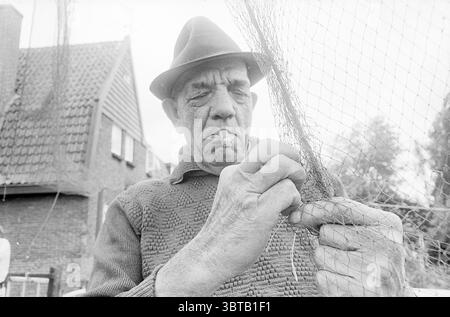 Pêcheur Jan Berk de Spaarndam finessing the nets Spaarndam, Whizgle News, Dutch Desk, pays-Bas, 1950 - 2000 on 21-06-1972. L'image montre ces rubriques. La scène montre un homme plus âgé engagé dans la tâche méticuleuse de réparer un filet de pêche. Son visage est altéré et marqué par les lignes de l'âge, véhiculant un sentiment d'expérience et de résilience. Il porte un chapeau de feutre sombre qui repose confortablement sur sa tête, son bord légèrement incliné. Ses mains, grandes et noueuses, saisissent habilement les fils délicats du filet, démontrant un contraste entre force et précision. Une cigarette pend de la sienne Banque D'Images