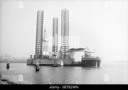 Super servant I avec plate-forme pétrolière dans IJmuiden Ships IJmuiden pays-Bas, Whizgle News, Dutch Desk, pays-Bas, 1950 - 2000 on 18-09-1982. Ces rubriques sont illustrées dans l'image. La scène présente un cadre maritime industriel caractérisé par une esthétique brutale et utilitaire. Au premier plan, une grande plate-forme de forage offshore se dresse bien en vue, surélevée sur des jambes hautes et minces qui s'étendent vers le haut comme des échafaudages géants. La plate-forme elle-même est peinte dans des tons discrets, principalement des gris et des blancs, contribuant à un aspect quelque peu austère contre son environnement. À côté de la plate-forme, un navire-citerne de taille Banque D'Images