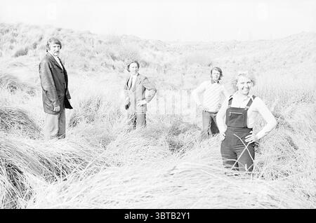 People in the dunes., Whizgle News, Dutch Desk, pays-Bas, 1950 - 2000. L'image montre ces rubriques. La scène montre quatre individus debout dans une zone luxuriante et herbeuse avec de grandes herbes se balançant doucement dans la brise. Chaque personne a une pose distincte, véhiculant un sentiment de confiance occasionnelle. La toile de fond présente un paysage vallonné, suggérant un cadre naturel qui s'étend au loin, avec des collines basses ou des dunes visibles. L'herbe est d'un vert doux, tandis que le filtrage de la lumière crée une qualité presque éthérée, donnant à la scène une sensation lumineuse et aérée. A gauche se trouve un homme dans un dar Banque D'Images