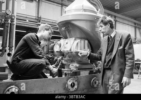 Lord of Deutekom Tekoma Company, Whizgle News, Dutch Desk, pays-Bas, 1950 - 2000 on 14-03-1991. Ce sont les éléments de l'image. La scène se déroule dans un environnement industriel spacieux, probablement un atelier ou une usine, caractérisé par une esthétique brute et utilitaire. L'arrière-plan est rempli de grandes machines et de structures métalliques, faisant allusion à une atmosphère de fabrication animée. Au premier plan, deux personnes participent à une discussion ciblée. Sur la gauche, un jeune homme s'agenouille à côté d'un grand équipement, vêtu d'une tenue de travail sombre et ajustée. Son expression suggère la concentration A. Banque D'Images