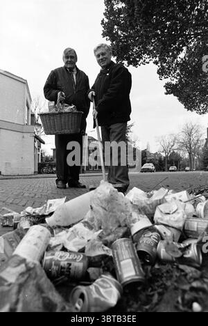 Deux hommes nettoient les ordures laissées par les étudiants Santpoort-Noord Kerkweg, Whizgle News, Dutch Desk, pays-Bas, 1950 - 2000 le 06-11-1990. L'image contient ces rubriques. La scène montre deux hommes plus âgés debout ensemble dans une rue, tous deux vêtus de vêtements sombres et décontractés. L'un tient un grand panier en osier, probablement utilisé pour ramasser la litière, tandis que l'autre s'appuie sur un bâton de marche. Leurs expressions semblent sincères et résolues, indiquant leur intention de nettoyer la zone. Au premier plan, il y a une pile dispersée de litière qui comprend diverses canettes vides, des papiers froissés et autres Banque D'Images