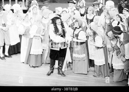 Velsen operetta performance Theater Opera operetta, Whizgle News, Dutch Desk, pays-Bas, 1950 - 2000 on 10-04-1986. L'image inclut ces rubriques. La scène se déroule sur une scène vibrante remplie d'un grand ensemble d'interprètes vêtus de costumes d'époque. Le décor suggère une ambiance festive, probablement issue d’une production historique ou théâtrale. À l'avant-garde, un personnage central se tient confiant, enfilant une tenue saisissante qui comprend un chapeau, un long manteau et des bottes brillantes, incarnant une présence dominante. Il semble s'engager avec animosité avec ceux qui l'entourent, faisant des gestes expressifs Banque D'Images